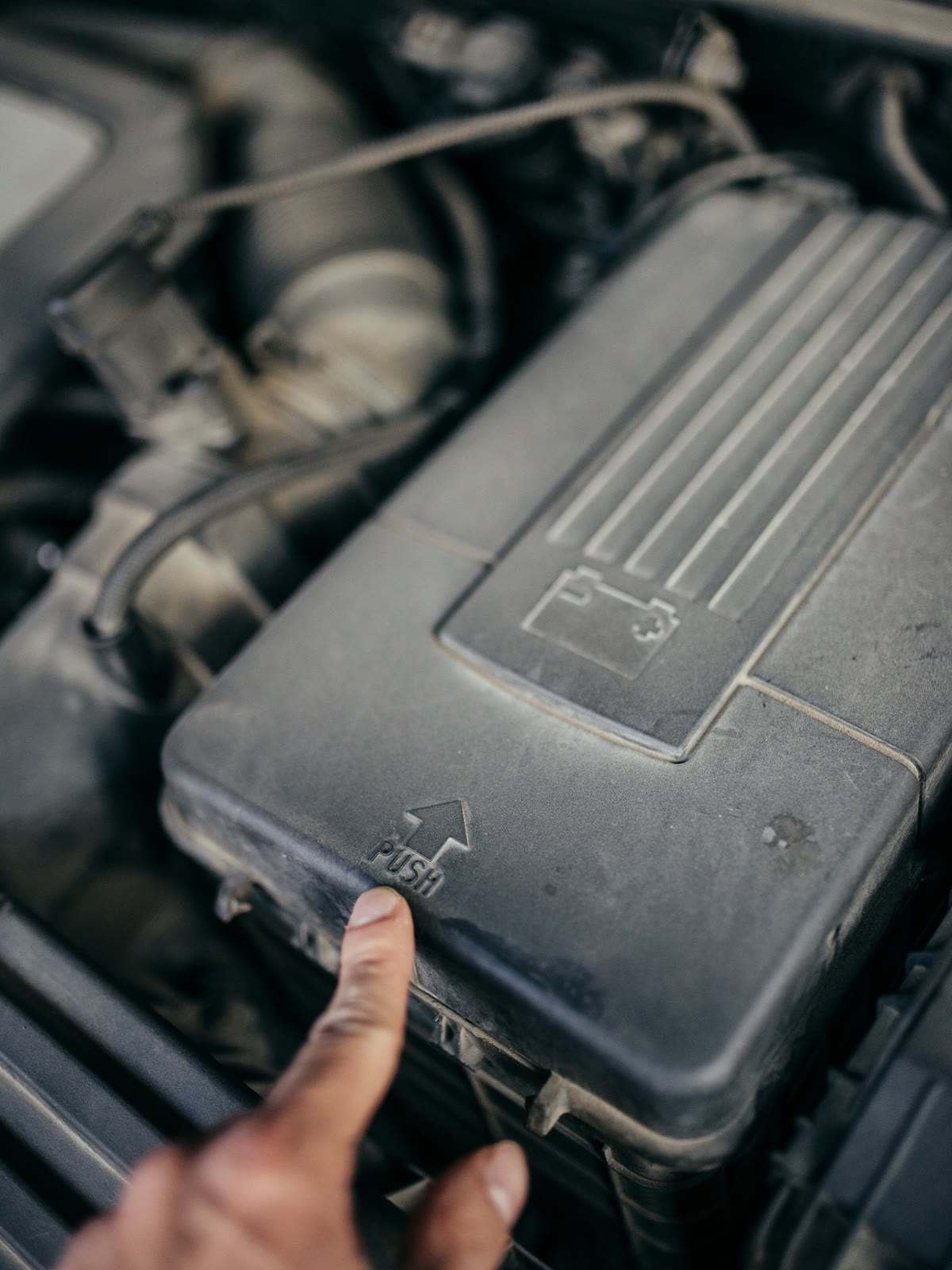 Mão apontando a tampa da bateria no motor de um carro, com o desenho da bateria gravado no plástico