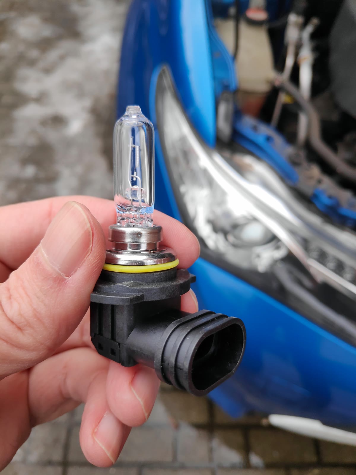Mão segurando uma lâmpada halógena de farol na frente de um carro azul com o farol aberto